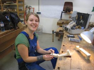 Junge Handwerkerin arbeitet an Holzteil am Werkbank, Werkzeuge im Hintergrund, fröhlich lächelnd.